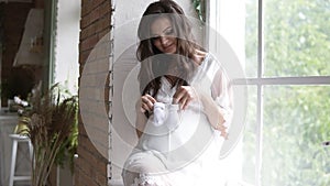 Pregnant, beautiful woman sitting on the sill by the window. Holding baby`s booties or socks, making little steps on her