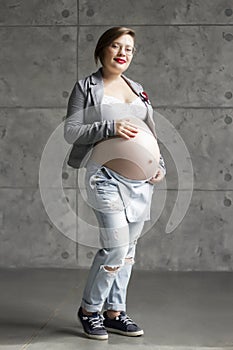 A pregnant beautiful young woman with glasses and red lipstick in a denim jumpsuit holds her stomach in a Studio against a gray wa
