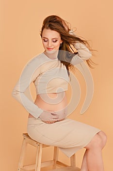 Pregnant beautiy girl in a long beige clothes on a beige background in the studio. Fashionable style for pregnant women. Studio