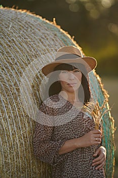 Pregnant beauty with black long hair leaned on a haystack, she was dressed in a fashionable dress, a straw hat on head