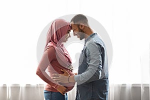 Pregnant Black Muslim Woman And Her Husband Hugging Near Window At Home