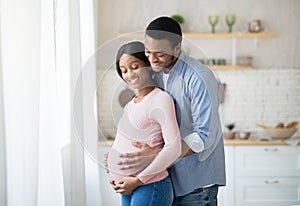 Pregnant black woman and her happy husband hugging next to window at home