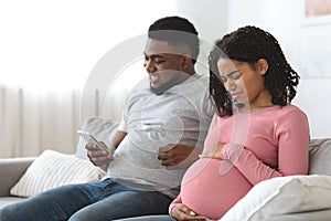 Pregnant black woman suffering from pain while husband using phone