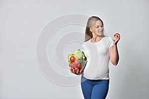 Pregnant blonde woman holding fresh vegetables in a bowl and tomato in her hand