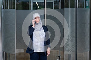 Pregnant business woman in hijab and suit talking on smartphone while leaving business center.