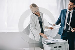 pregnant businesswoman talking on smartphone while using printer and businessman with papers standing behind