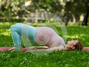 Pregnant caucasian woman doing yoga in the park. Prenatal shoulder bridge exercise.