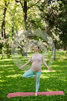 Pregnant caucasian woman doing yoga in the park. Tree pose.