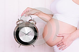 Pregnant caucasian woman is holding big clock close to her tummy