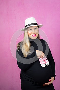 Pregnant Caucasian woman posing holding baby shoes for a maternity shoot on a pink background