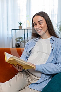 Pregnant Caucasian woman reading book advice child care guide feeling relax on cozy couch at home