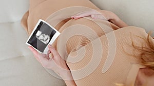 Pregnant caucasian woman resting at home. A young expectant mother looks at ultrasound pictures of her baby