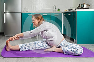 A pregnant Caucasian woman is sitting and doing yoga, making leans to the left side, setting her leg aside. The kitchen is in the
