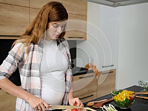 Pregnant Caucasian woman slicing tomatoes on a cutting board,preparing delicious healthy meal. A bow with a colorful healthy salad