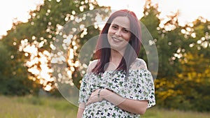 Pregnant caucasian woman in summer dress posing outdoors. Close up of young lady holding two hands on her pregnant belly