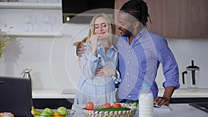 Pregnant Caucasian woman tasting organic ingredients for healthful salad as African American man hugging spouse talking
