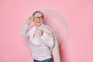 Pregnant cheerful ethnic woman smiling looking aside, putting trendy glasses on isolated pink background. Copy ad space