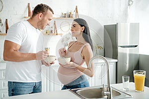 Pregnant couple eating cereals with milk from bowls in kitchen