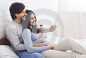 Pregnant couple relaxing with smartphone on bed at home