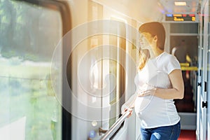 Pregnant european woman is staying and holding her own tummy close to train window while she traveling by rail way and holding han