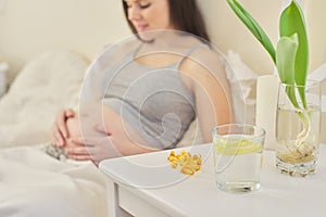 Pregnant female in home interior sitting in bed, vitamin capsules close up