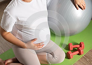 A pregnant girl with a big belly is engaged with dumbbells against the background of a gray fitball ball. Sports activities in the