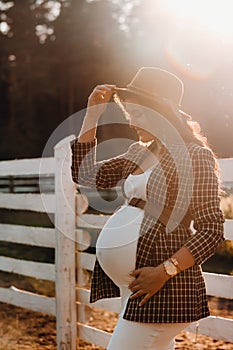 A pregnant girl with a big belly in a hat near a horse corral in nature at sunset.Stylish pregnant woman in a brown dress with
