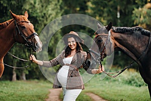 Pregnant girl with a big belly in a hat next to horses in the forest in nature.Stylish pregnant woman in the brown dress with the