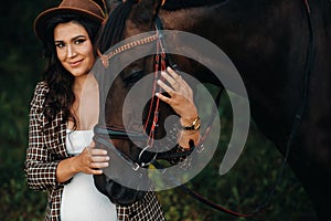 Pregnant girl with a big belly in a hat next to horses in the forest in nature.Stylish pregnant woman in the brown dress with the