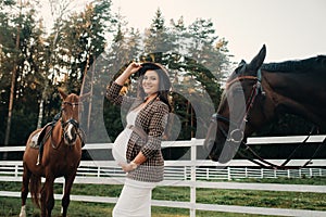 A pregnant girl with a big belly in a hat next to horses near a paddock in nature.Stylish pregnant woman in a brown dress with