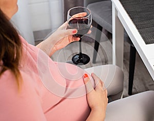 A pregnant girl with a big belly in a pink blouse holds a glass of wine. The concept of drinking alcohol during pregnancy, a bad