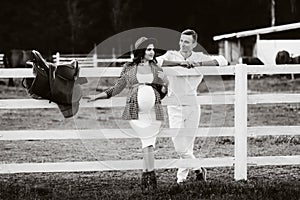 A pregnant girl in a hat and her husband in white clothes stand next to the horse corral.a stylish couple waiting for a child