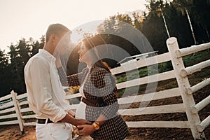 A pregnant girl in a hat and her husband in white clothes stand next to a horse corral at sunset.a stylish couple is waiting for a