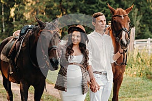 A pregnant girl in a hat and her husband in white clothes stand next to horses in the forest in nature.Stylish pregnant woman with