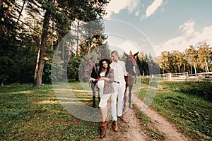 A pregnant girl in a hat and her husband in white clothes stand next to horses in the forest in nature.Stylish pregnant woman with
