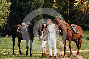 A pregnant girl in a hat and her husband in white clothes stand next to horses in the forest in nature.Stylish pregnant woman with