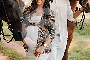 A pregnant girl in a hat and her husband in white clothes stand next to horses in the forest in nature.Stylish pregnant woman with