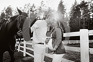 A pregnant girl in a hat and her husband in white clothes stand next to the horses near the horse corral at sunset.Stylish