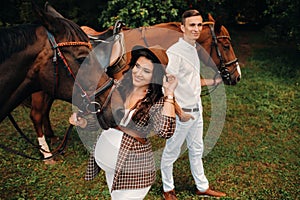 A pregnant girl in a hat and a man in white clothes stand next to horses in the forest in nature.Stylish pregnant woman with her