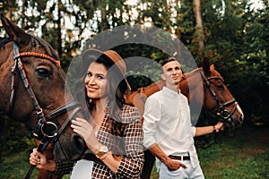 A pregnant girl in a hat and a man in white clothes stand next to horses in the forest in nature.Stylish pregnant woman with her