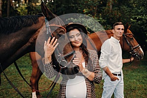 A pregnant girl in a hat and a man in white clothes stand next to horses in the forest in nature.Stylish pregnant woman with her