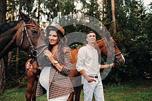 A pregnant girl in a hat and a man in white clothes stand next to horses in the forest in nature.Stylish pregnant woman with her