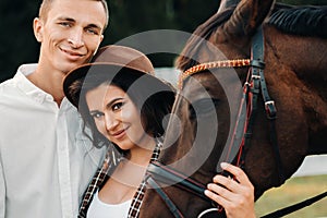 A pregnant girl in a hat and a man in white clothes stand next to horses near a white fence.Stylish pregnant woman with a man with