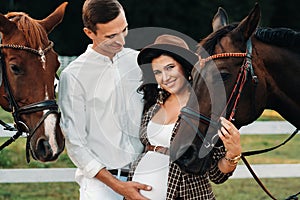 A pregnant girl in a hat and a man in white clothes stand next to horses near a white fence.Stylish pregnant woman with a man with