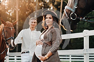 A pregnant girl in a hat and a man in white clothes stand next to horses near a white fence.Stylish pregnant woman with a man with