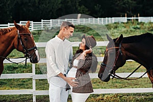 A pregnant girl in a hat and a man in white clothes stand next to horses near a white fence.Stylish pregnant woman with a man with