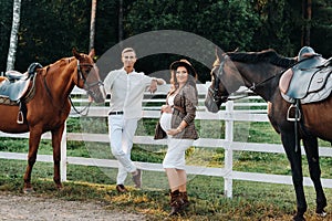 A pregnant girl in a hat and a man in white clothes stand next to horses near a white fence.Stylish pregnant woman with a man with