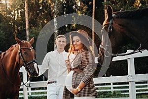 A pregnant girl in a hat and a man in white clothes stand next to horses near a white fence.Stylish pregnant woman with a man with