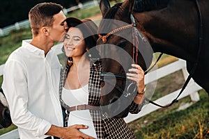 A pregnant girl in a hat and a man in white clothes stand next to horses near a white fence.Stylish pregnant woman with a man with