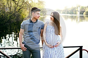 A pregnant girl and her boyfriend are sitting on the pier and looking at each other, a beautiful river and sunset